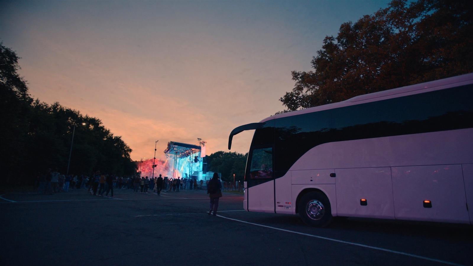 Autocar de luxe garé près de l'entrée d'un festival de musique au crépuscule, scène principale illuminée en arrière-plan